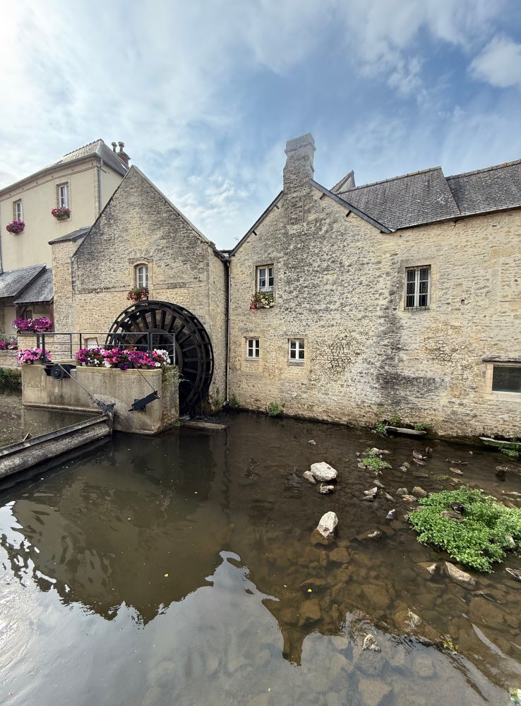 Frankreich, Flussbett, im Hintergrund Häuser, ein Mühlrad, Blumenkästen mit roten, weißen, rosa Blumen. 