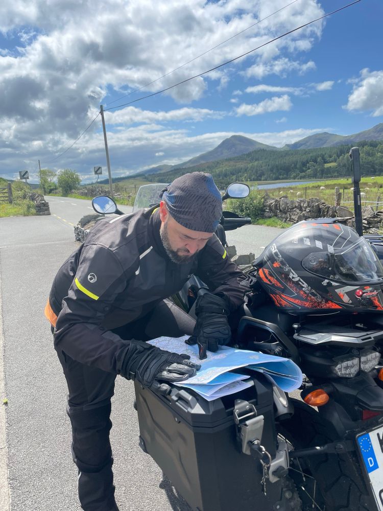 Der Motorradfahren beugt sich an seinem Motorrad über ein Landkarte, sein Helm sowie der Helm der Sozia liegen auf der Maschine, im Hintergrund blauer Himmel, Wiesen und Gebirge. 