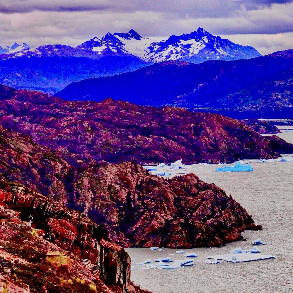 A jagged tundra lined shoreline along the glacial milk waters of Gray Lake. A bright blue ice berg floats in a bay. 
Snowcapped peaks on the horizon under a cloud filled sky.  