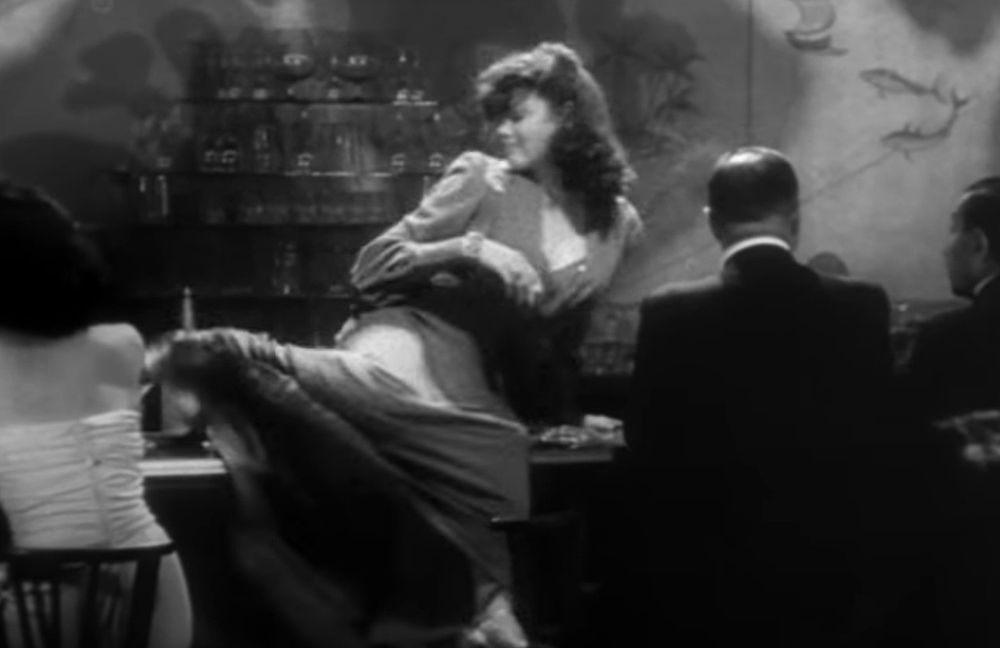 Gene, hair tousled, obviously drunk, perched on a bar and lifting her leg up to kick stuff off it