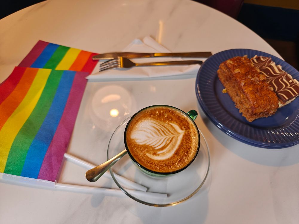 White table with a flat white in a fancy cup, 2 pieces of cake, and two small rainbow pride flags