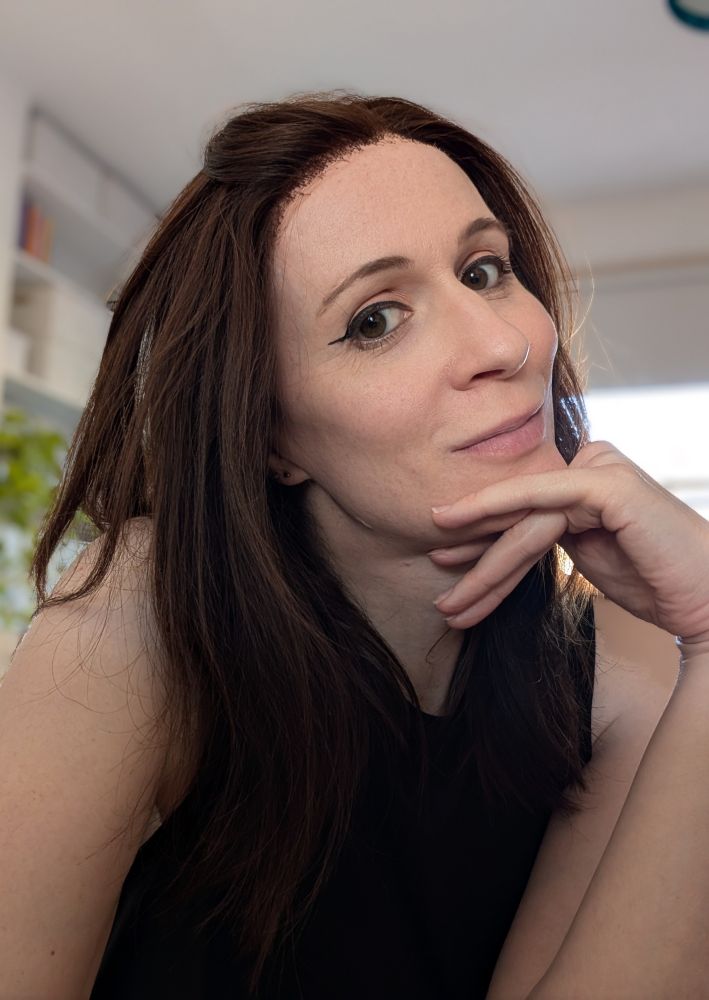 Close up self portrait of the artist, dressed as before. Cropped below the chest, upper right arm visible, left arm bent at the elbow so my hand touches my chin, hair pushed back from my face, winged eyeliner on display, eyes looking slightly downwards into the camera, and what has recently seen described by multiple, unrelated, people as an enigmatic smile on my face 