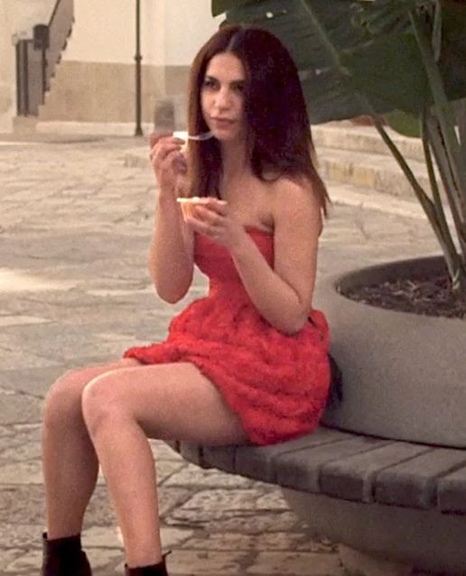 A still from Spring (2014), showing Nadia Hilker, wearing a strapless red mini dress and black ankle boots, long dark brown hair worn down and falling onto her shoulders, sitting on a bench eating ice cream in a picturesque Italian town square