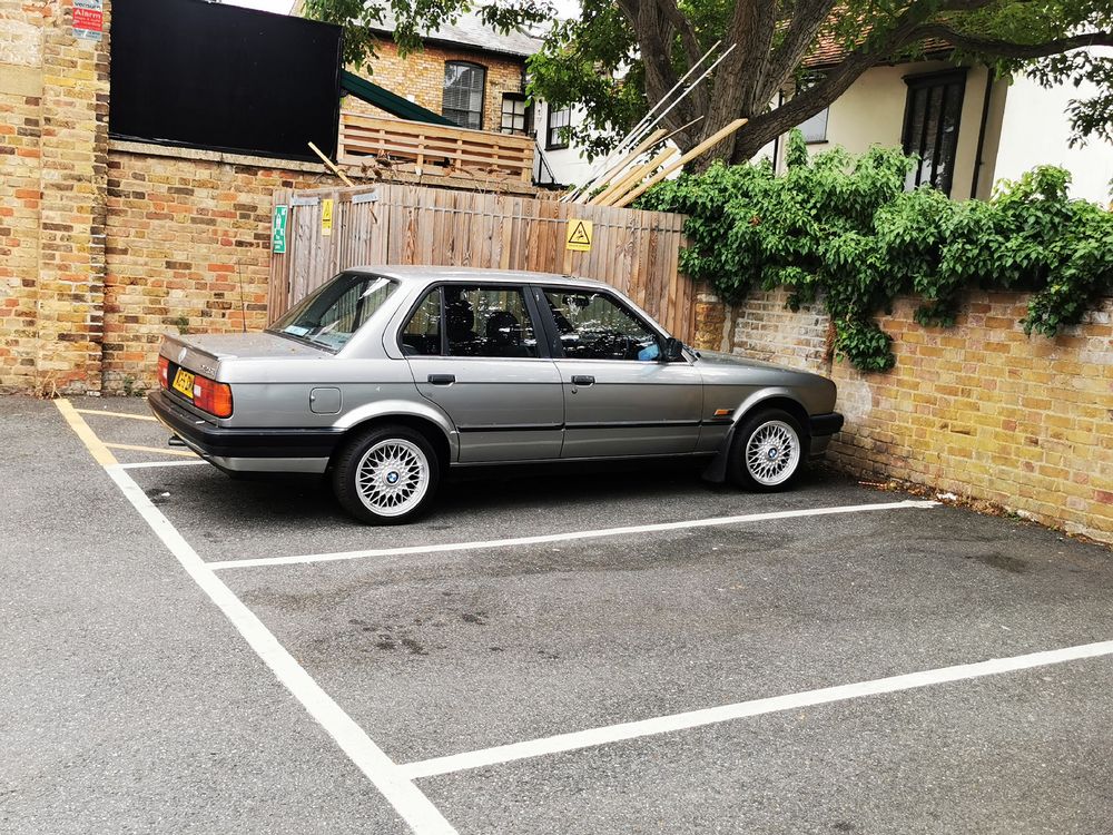 A silver late 90s BMW 3 SERIES four door salon parked in a small car park. The car is silver grey with period alloy wheels. 