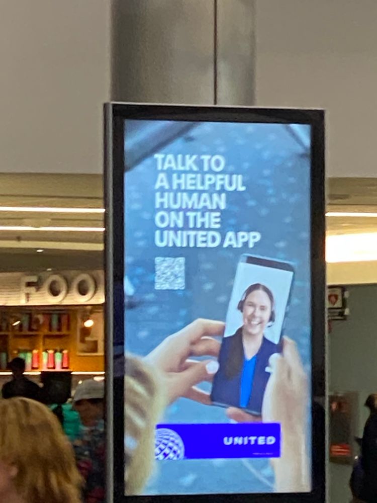 Electronic airport advertising stating “Talk to a helpful human on the United App” with a picture of someone holding a phone with an image of a smiling United employee. 