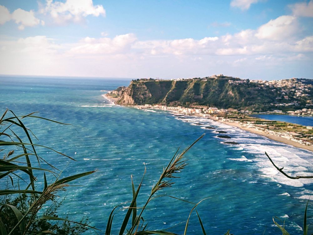 The Naples coastline in Italy shows grass in the foreground then behind that is blue green water hitting the shores with a white foam, you can see many small white structures lining the beach. Further back in the background you see a large green and dark sandy colored cliff with some small structures sprinkled across it's flat top, more inland on the cliff is a large cluster of white and redtan structures. The weather is slightly cloudy and has a light  blue sky .