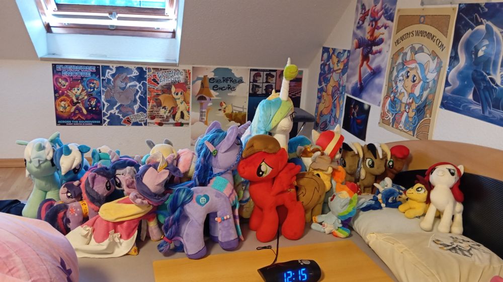 A colorful collection of plush ponies on a bed, a number of pony convention posters on the walls.