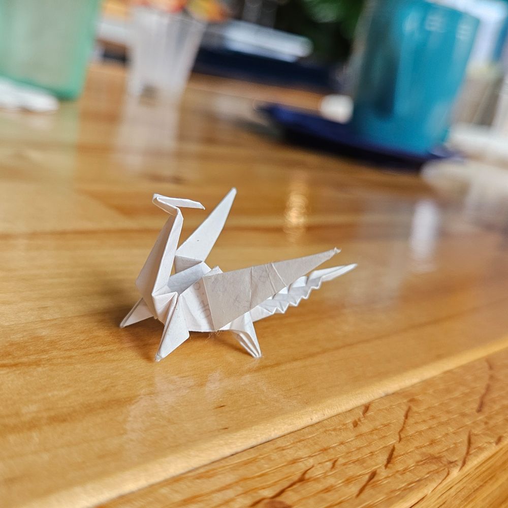 Tiny origami dragon sitting on a bar countertop with a blurry background of cups and mugs