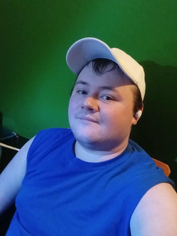 Selfie of a young white man in a blue cutoff tank and white baseball cap. He has wireless earbuds in.