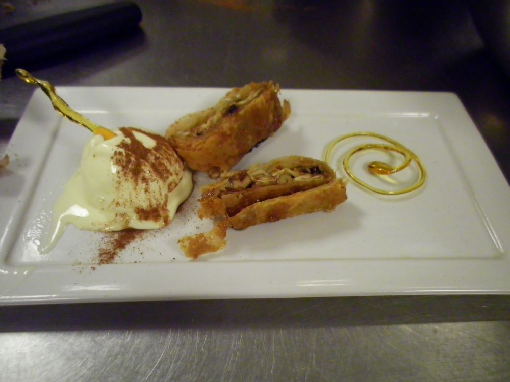 A rectangular plate. In the middle, there are 2 small slices of apple streudel. On the left is a scoop of vanilla ice cream with a dusting of cinnamon, and a freeform caramel shape. On the right is a spiral caramel shape