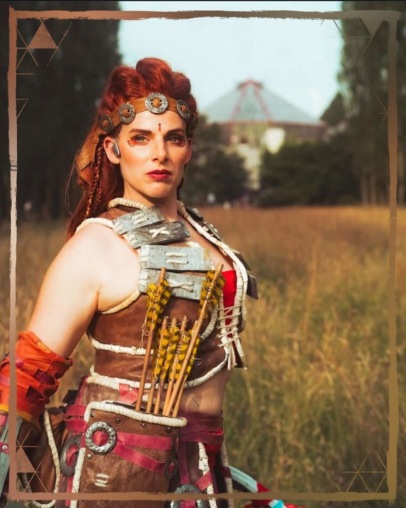 A photo of an Aloy cosplayer wearing Oseram armor, standing proudly while holding her spear in one hand, down by her side.