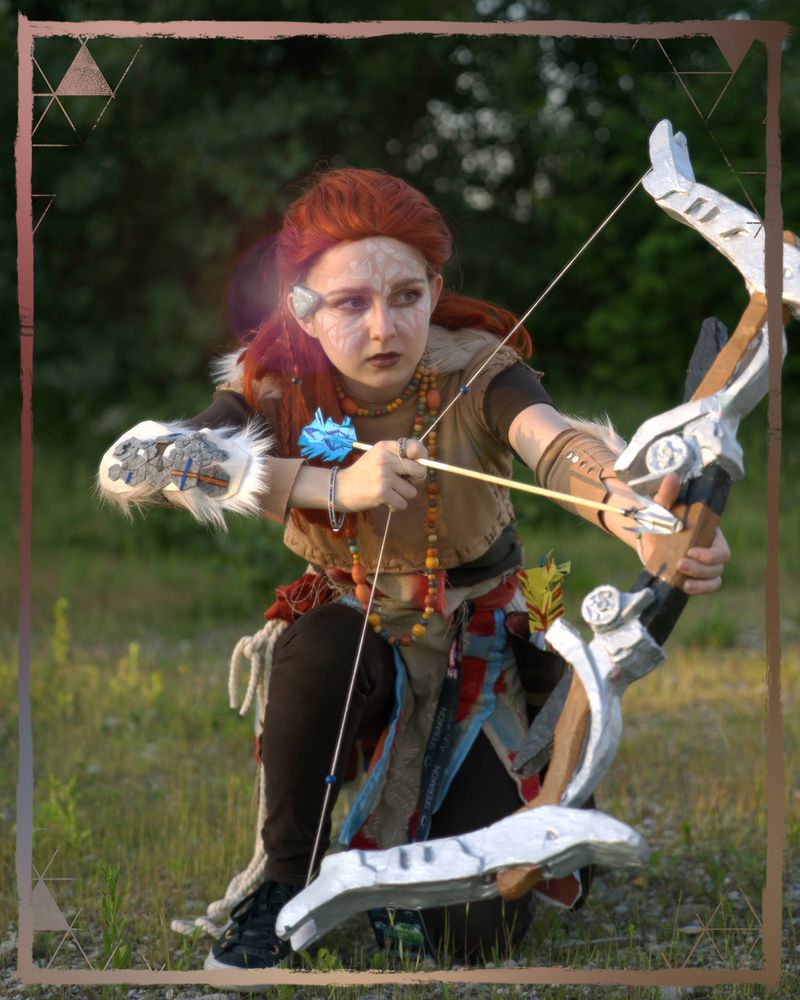 A photo of an Aloy cosplayer kneeling down with her bow drawn in front of her. She is also wearing white Utaru face paint.