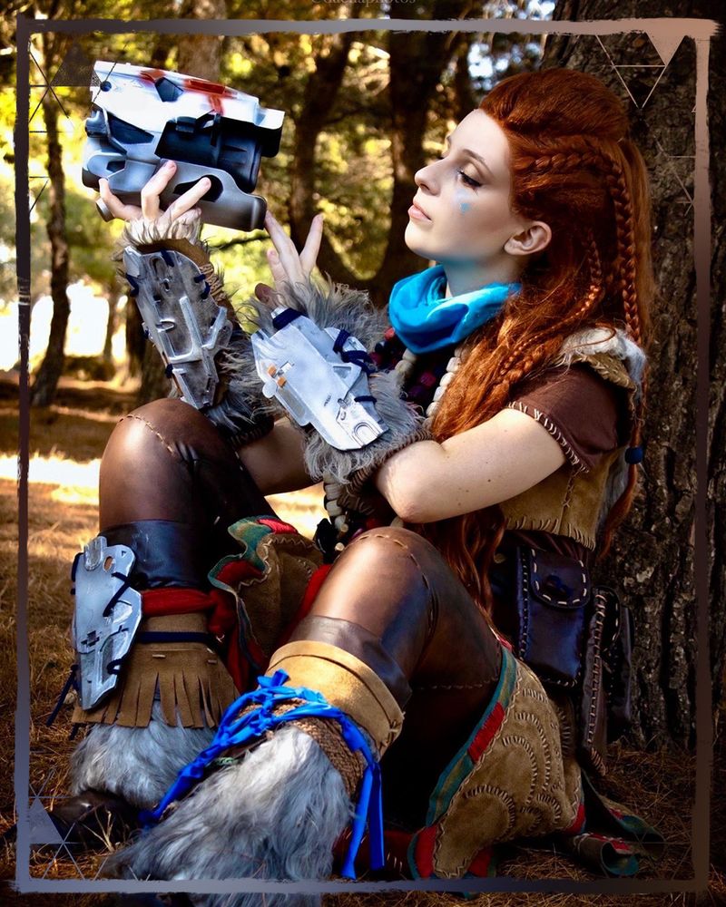 A photo of an Aloy cosplayer sitting by a tree. She is holding a Watcher machine's head in her hands as she inspects it.