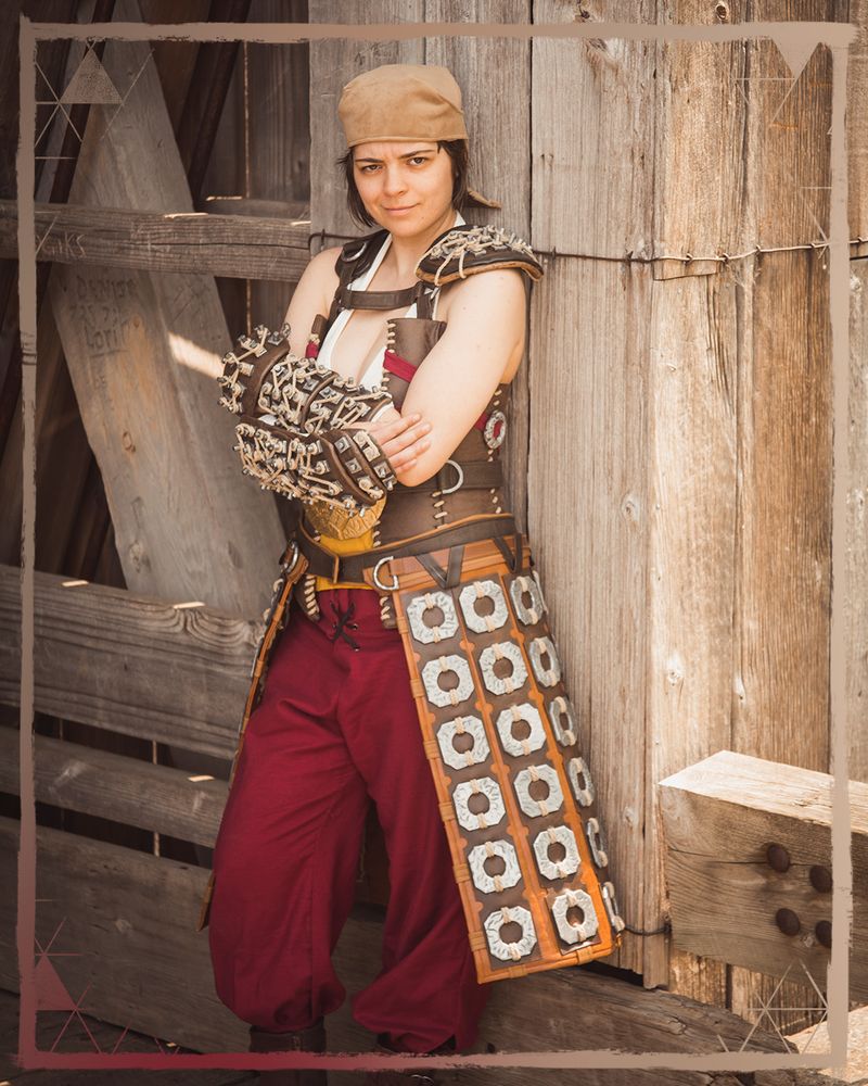 A photo of a Petra cosplayer standing against a wooden wall with her arms folded, looking directly at the camera.