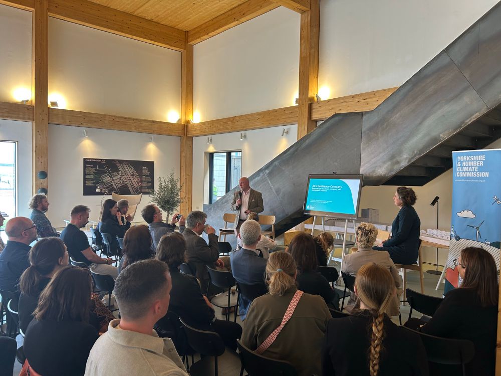People are seated in rows of chairs listening to a male speaker in a timber framed room
with white walls. He is standing at the front giving a PowerPoint presentation on a large screen.