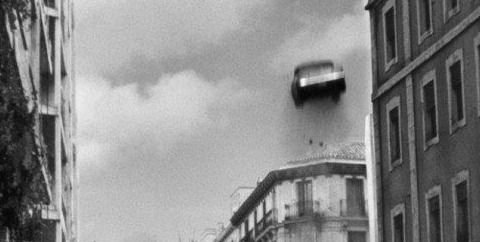 Black and white picture. The tops of buildings can be seen from a civilian's perspective. A car can be seen in the air near the rooftops, with motion blur indicating an upward motion. There is some rubble and dust underneath the car.