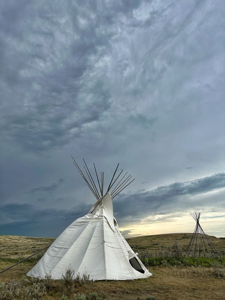 The dark clouds of an encroaching storm sweeps across the sky over the prairie. A white tipi blows in the heavy winds.