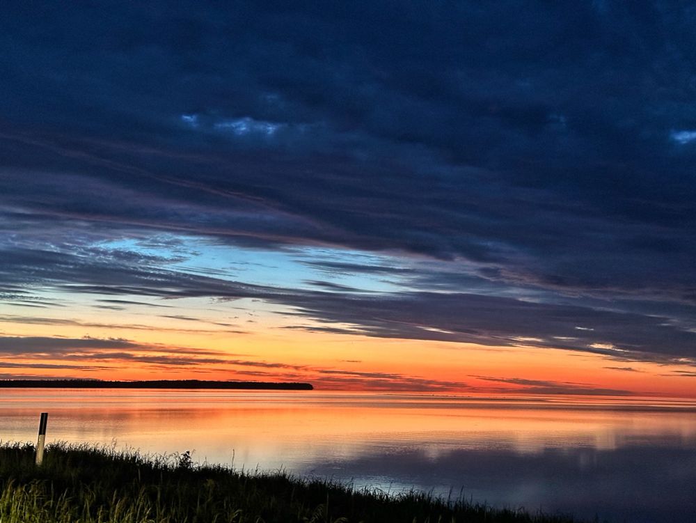 A wedge of orange sky glows above the horizon and is reflected in the Still waters of Lake Winnipeg at sunrise. Slate gray clouds fill the rest of the sky at early morning.