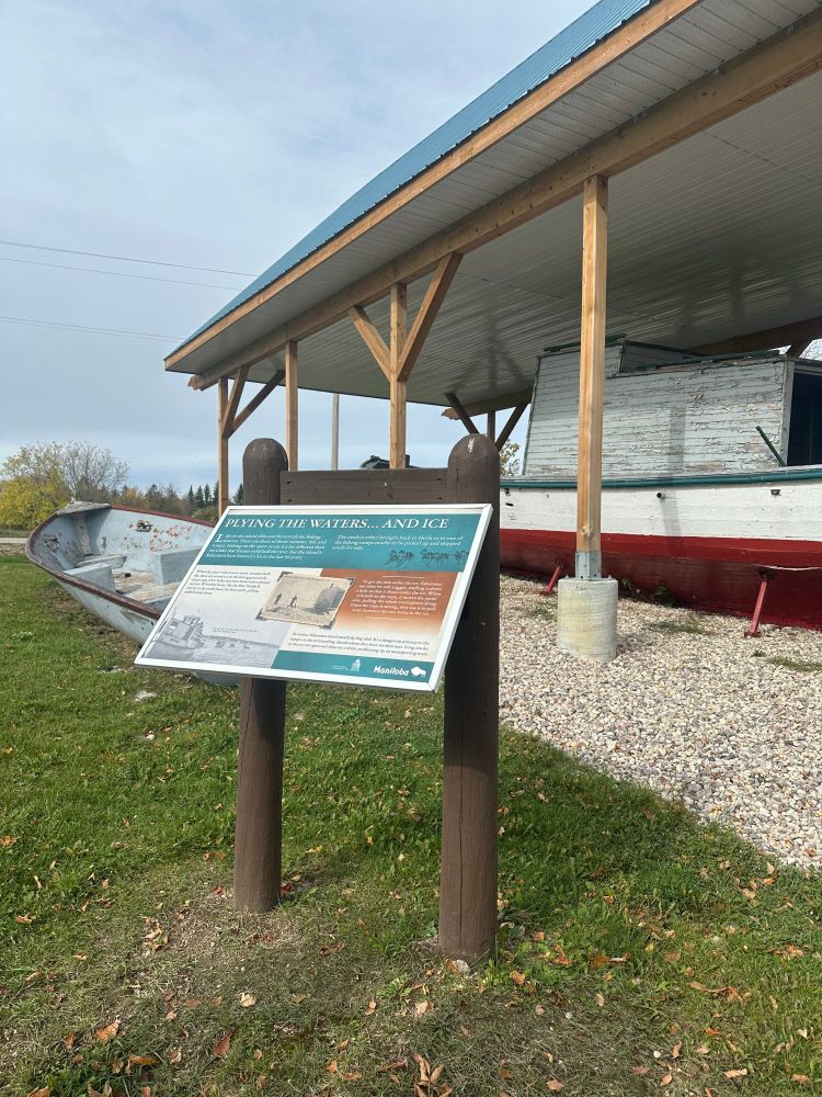 A blue-topped interpretive panel titled - Plying the Water and Ice sits in front of an old wooded whitefish boat protected by the elements by a large roofed structure. The sign tells the story of the start of the commercial fishing industry on Lake Winnipeg.