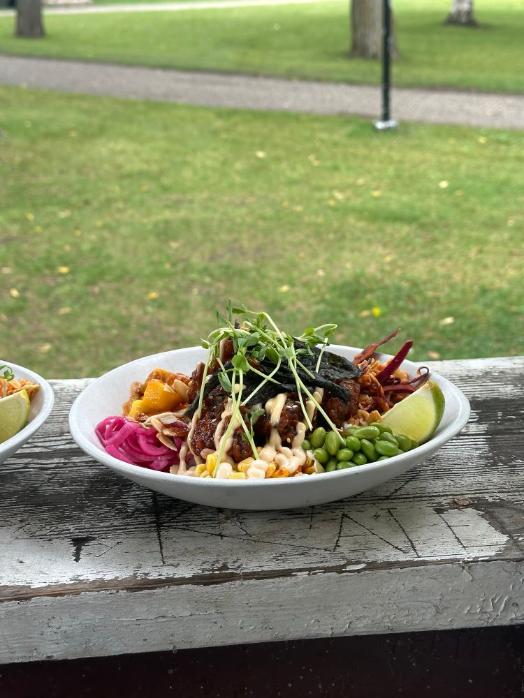 A bowl of karage chicken with corn, edamame, mango, pickled onions, lime, and rice sits on a wooden ledge outside.