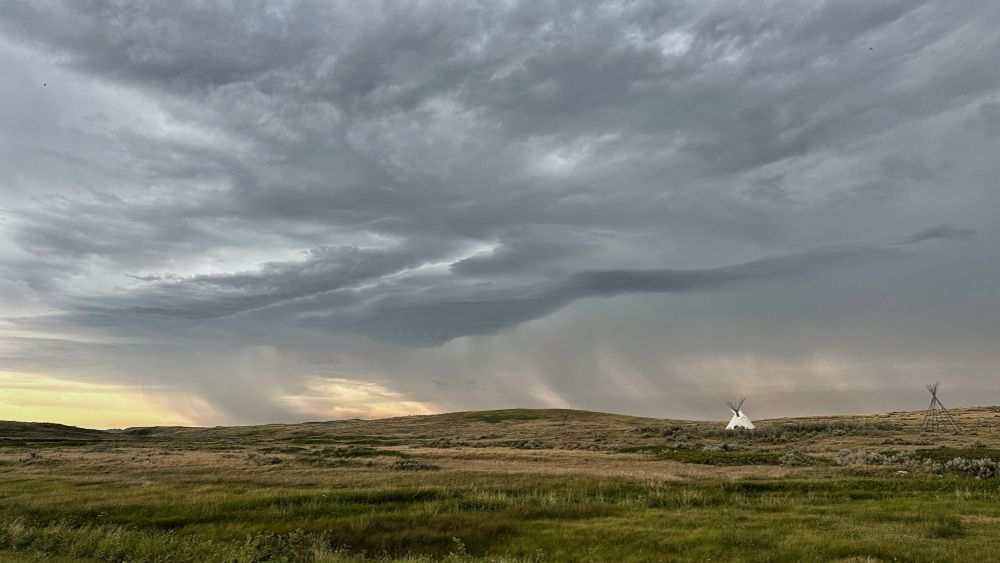Dark thunder, clouds sweep over the open prairie, dropping curtains of rain in the distance over the pale orange of the early morning sky. A white tipi contrast sharply against the pale green grasses, and the oncoming storm.