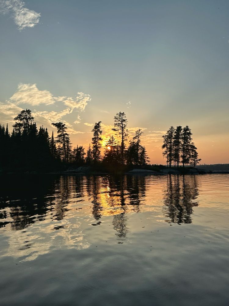 The pale yellow sun disappears behind a collection of tall pine trees in late evening. The sky and trees are reflected in the mostly Stillwater in the foreground.
