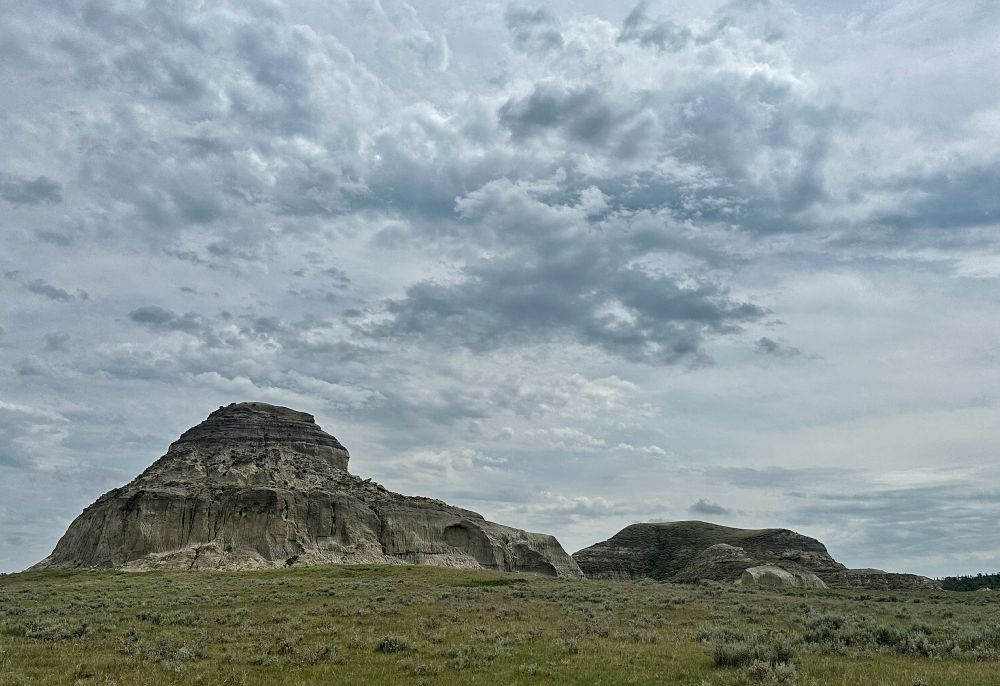 A view of the same butte from another side reveals a longer foot off to one end and steep walls around the base. Clouds add drama to the photo. 