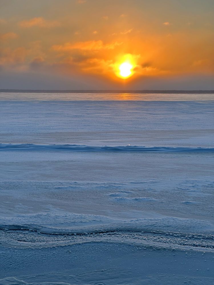 Looking out over frozen lake, the orange ball of the sun is just clearing the low clouds that skirt the horizon. The ice is purple blue with very little to mar the surface except for a few small ridges that are starting to form.