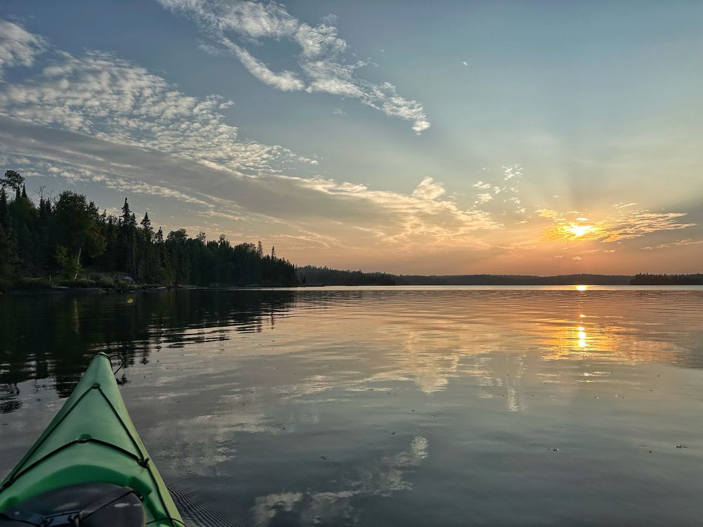 The sun disappearing behind a small bank of clouds is reflected in the Stillwaters of a boreal lake. You can see the bow of a green kayak in the foreground on the left of the image pointing towards the tree lined shore.