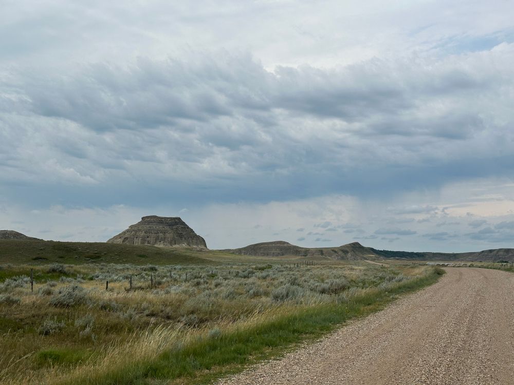 A large sandstone butte looms over the prairie as a lonely gravel road curves towards it in the foreground. Clouds swirl in the sky above it. 