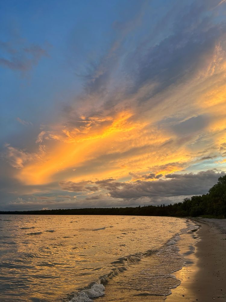 Ablaze of orange cloud sweeps diagonally across a turbulent sky. The colour reflects dully on the wavy lake in the foreground and shines in the water left behind on the sand.