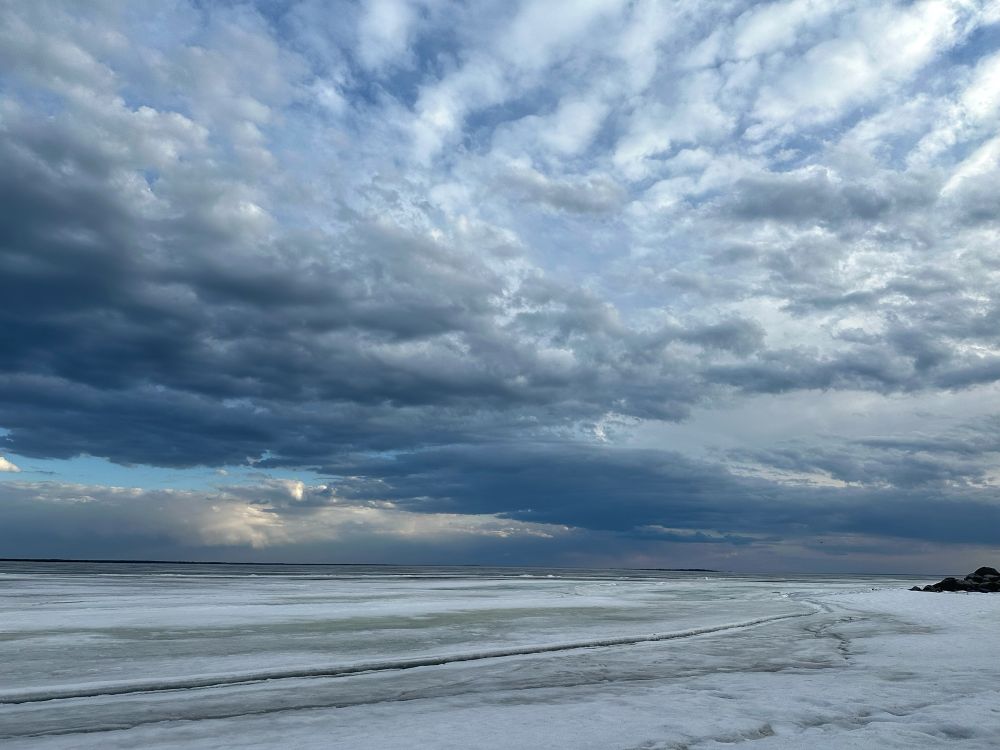 Looking out over a frozen lake, where the ice is starting to rot. There are patches of dark ice all over the place showing where it’s getting thin. The sky is filled with clouds that are lit up by the sun, making everything look silvery