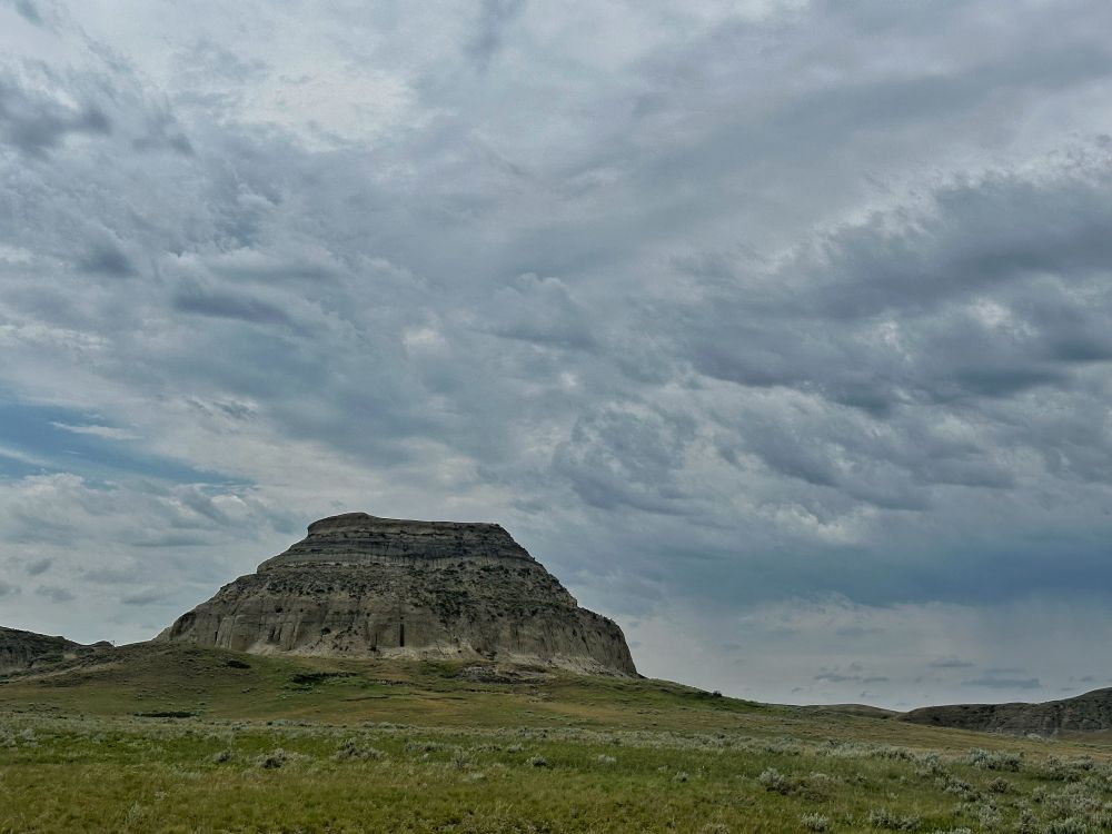 A massive sandstone butte stands sentinel over the grassland, stark against the clouds looming above it. 