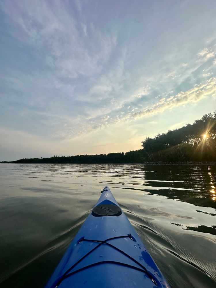 A wide-angle shot across the bow of a blue kayak looking over a mostly still lake, the setting sun shines through the trees on the shore along the right, lighting up the thin clouds overhead with a golden glow. 