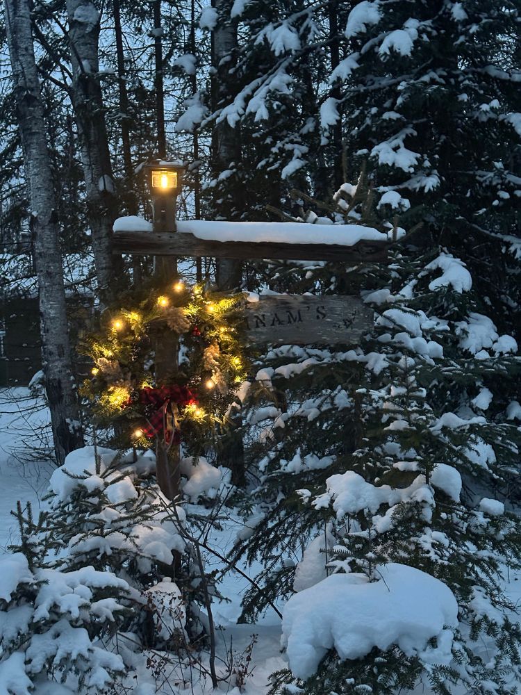 A wreath made of spruce and fur boughs with a red plaid bow and small lights hangs on the post that is holding a cottage sign surrounded by snow covered spruce trees. On top of the post is a small solar light that glows in the early evening gloom.