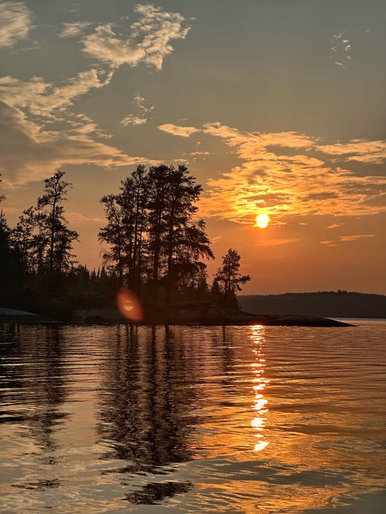 The orange Sun reflects off the mostly Stillwater as it sinks down over a distant boreal shore. In the foreground, a small island is silhouette by the bright orange light. It’s tall pine trees standing dark against the evening light.