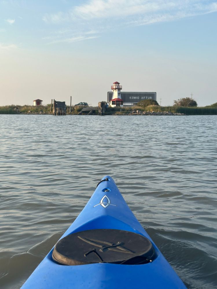 Looking over the bow of a blue kayak across a slightly wavy lake towards a sign with a large white and red lighthouse built into the left side of it. The sign says Komid aftur which I think means come again in Icelandic.