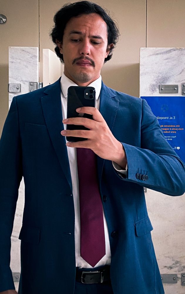 Homem caucasiano de terno azul e gravata vinho segurando o celular com a mão direita de frente ao espelho