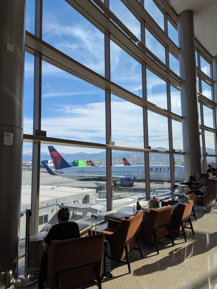 View from the SLC airport lobby windows that stretch floor to ceiling. Chairs are lined in front of the windows. A series of Delta airplanes are visible with a mountain backdrop.