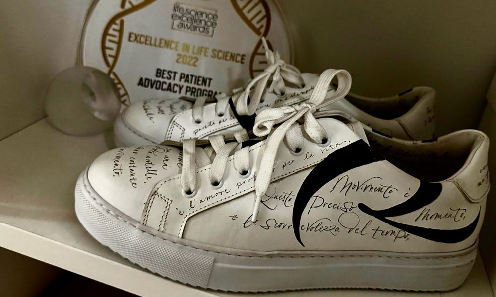 A pair of white sneakers with black calligraphy-style writing on them sit on a white shelf. The shoes feature handwritten messages in Italian about movement, life, and multiple sclerosis. Behind them, a golden and white award plaque reads “Excellence in Life Science 2022 – Best Patient Advocacy Program.”
