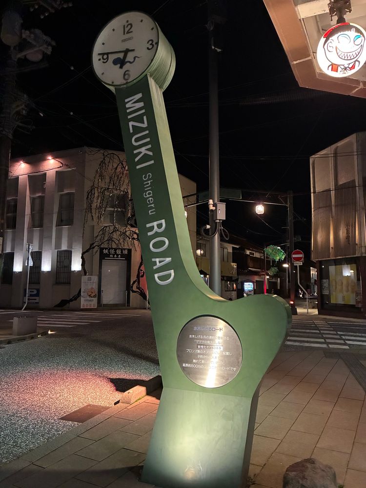 時計台と、夜の街の風景。時計台には「MIZUKI Shigeru ROAD」と書かれている。
