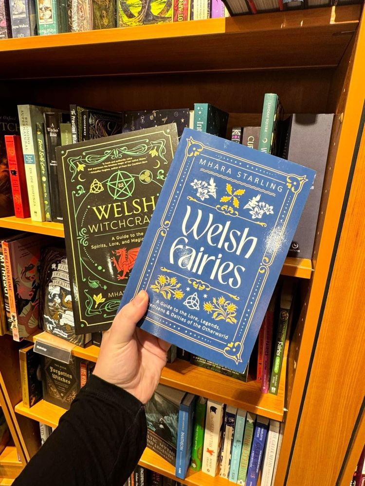 A hand holding two books in a bookshop - the first book on the left is ‘Welsh Witchcraft’ by Mhara Starling. It has a black cover with ornate green and yellow designs and a large red dragon at the centre. The second book is ‘Welsh Fairies’ also by Mhara Starling, this book has a blue cover with ornate white and yellow designs. 