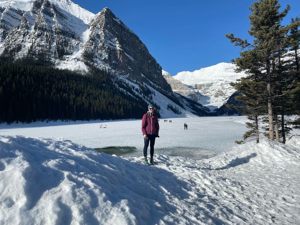 Person in a purple jacked standing in a snowy environment in front of a frozen lake. Mountains, trees and blue sky in the background.