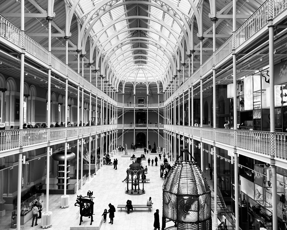 Black & white photo depicting an interior view of the National Museum of Scotland in Edinburgh’s Chambers Street. The Victorian architecture is grand, like a giant glasshouse with sweeping arches, its design inspired by the Crystal Palace in London. There are visitors of all ages wandering around, examining the museum’s many & varied artefacts.