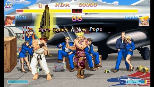 Screenshot from Street Fighter 2 of Ryu fighting Guile on Guile's stage, the tarmac. Ryu is wearing a photo-edited tall hat with a yellow glow. The words "Here Comes A New Pope" are on the screen.