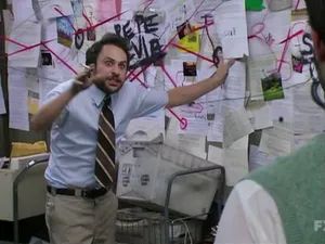 The image depicts a man in a disheveled office setting, surrounded by walls covered in papers, notes, and graffiti. He has a somewhat frantic expression and is gesturing with his arms outstretched, as if trying to make sense of the chaotic environment around him. The man is wearing a white dress shirt, tie, and slacks, suggesting he is meant to portray someone working in an office or professional setting that has descended into disarray. The overall scene conveys a sense of confusion, conspiracy theories, and an overwhelming amount of disorganized information or ideas.