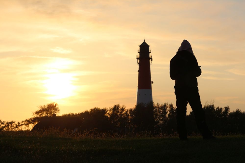 Sonnenuntergang auf der Insel Poel. Man sieht im Vordergrund den Deich der Insel auf der rechten Seite steht eine Person links daneben der Leuchtturm der Insel dazwischen einige Bäume. Die Sonne strahlt im Hintergrund in einem tiefen orange
