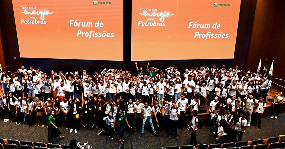 A imagem mostra um grande grupo de pessoas reunidas em um auditório. Elas estão vestindo camisetas brancas e parecem animadas, muitas delas estão acenando e sorrindo. Ao fundo, há dois telões exibindo as palavras "Fórum de Profissões" e "Traga a sua energia para a Petrobras".