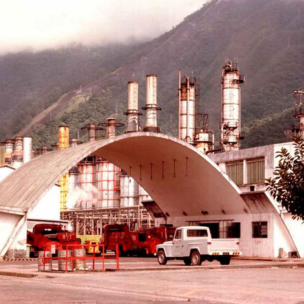 Imagem de uma instalação industrial com chaminés altas ao fundo, sob um céu nublado. Em primeiro plano, há um pórtico curvo e veículos, incluindo caminhões vermelhos e um carro branco. Montanhas verdes se erguem ao fundo.