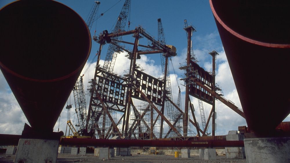 A imagem mostra uma estrutura industrial em construção, com várias gruas e peças de metal expostas. No primeiro plano, há um tubo grande com a abertura voltada para a câmera. Ao fundo, são visíveis estruturas metálicas de grande porte, possivelmente parte de uma plataforma ou construção similar, com scaffolding (andaimes) ao redor.